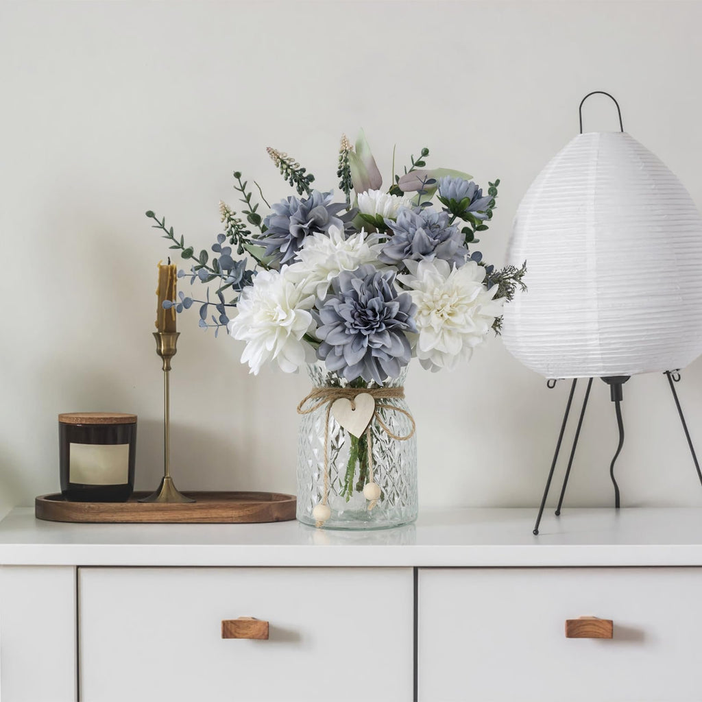 Blue and white artificial dahlia flowers in a glass vase with twine and wooden heart decoration, next to a candle and a lamp.