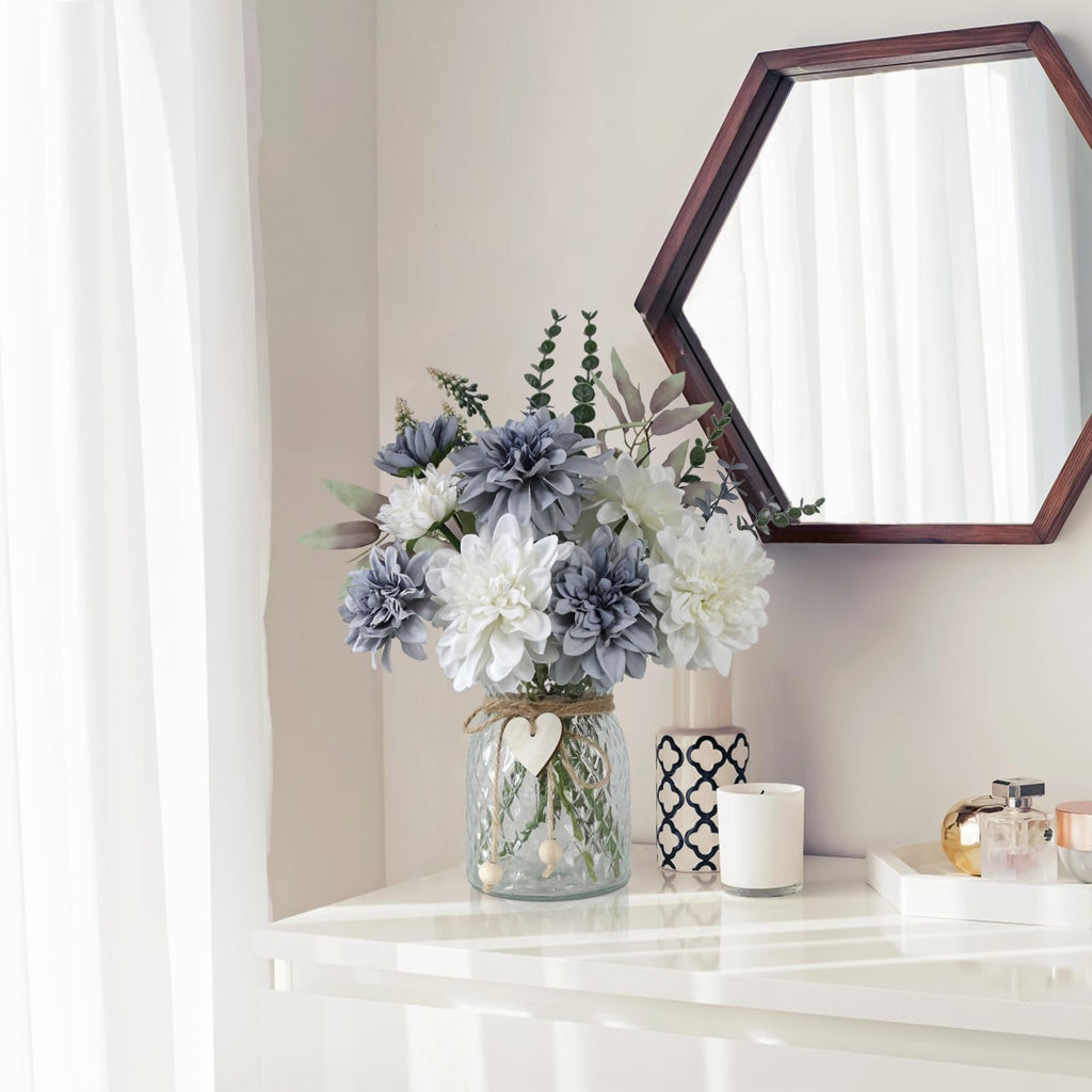 Blue and white artificial dahlia flowers in a clear glass vase with twine and wooden heart accent, on a white shelf with decorative items.