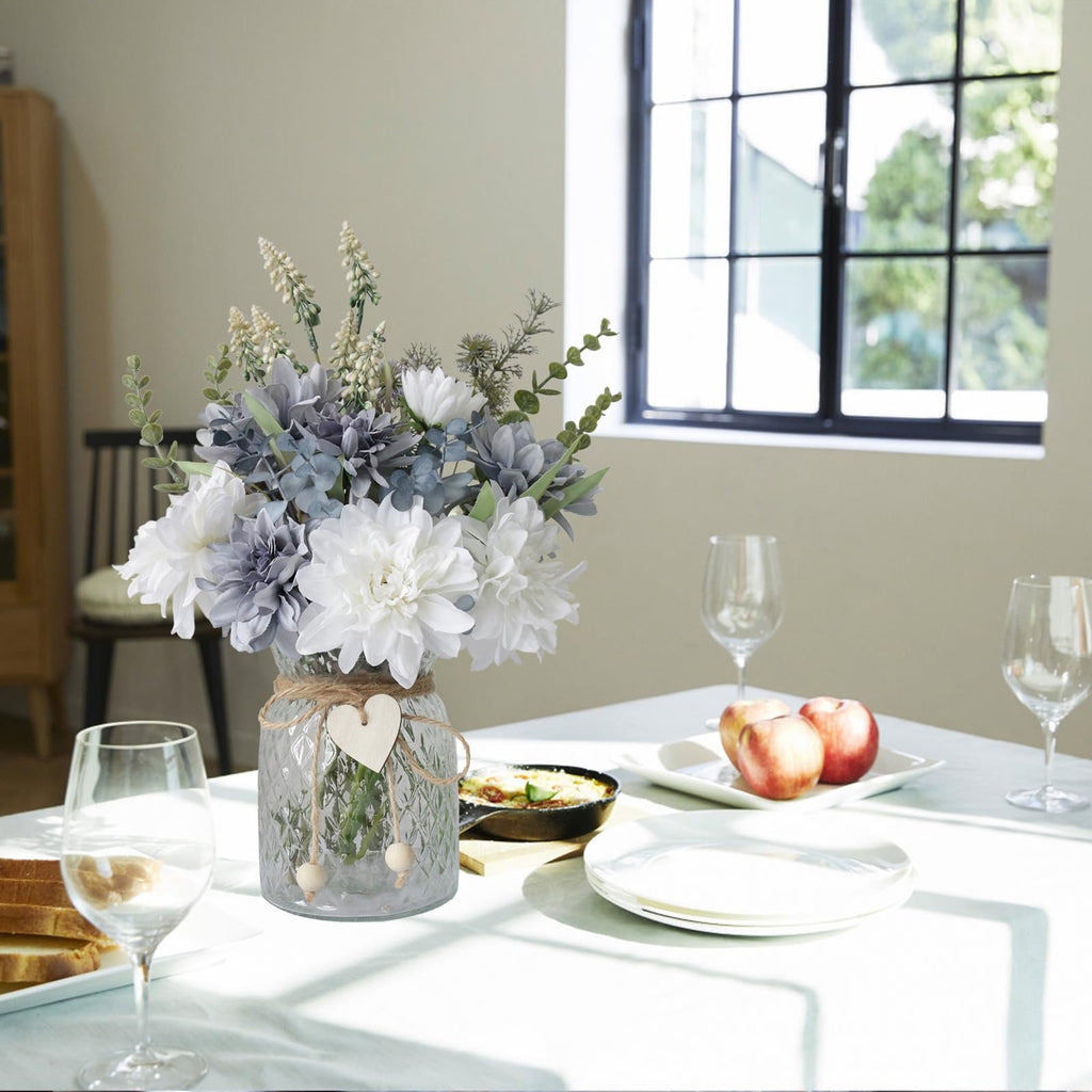 Artificial dahlia flower arrangement in a textured glass vase on a table setting with plates and wine glasses.