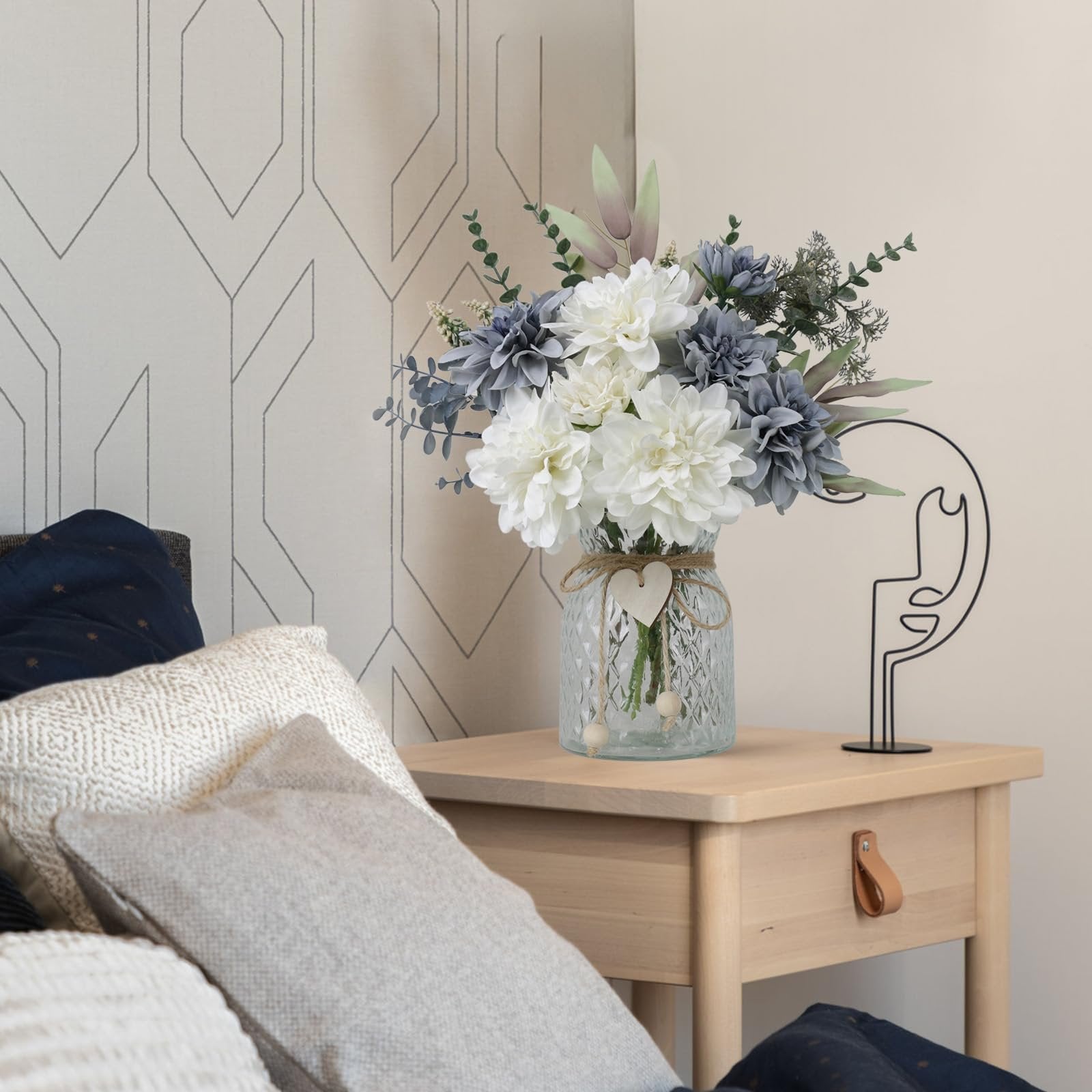 Artificial dahlia floral arrangement in a glass vase on a wooden nightstand with pillows.