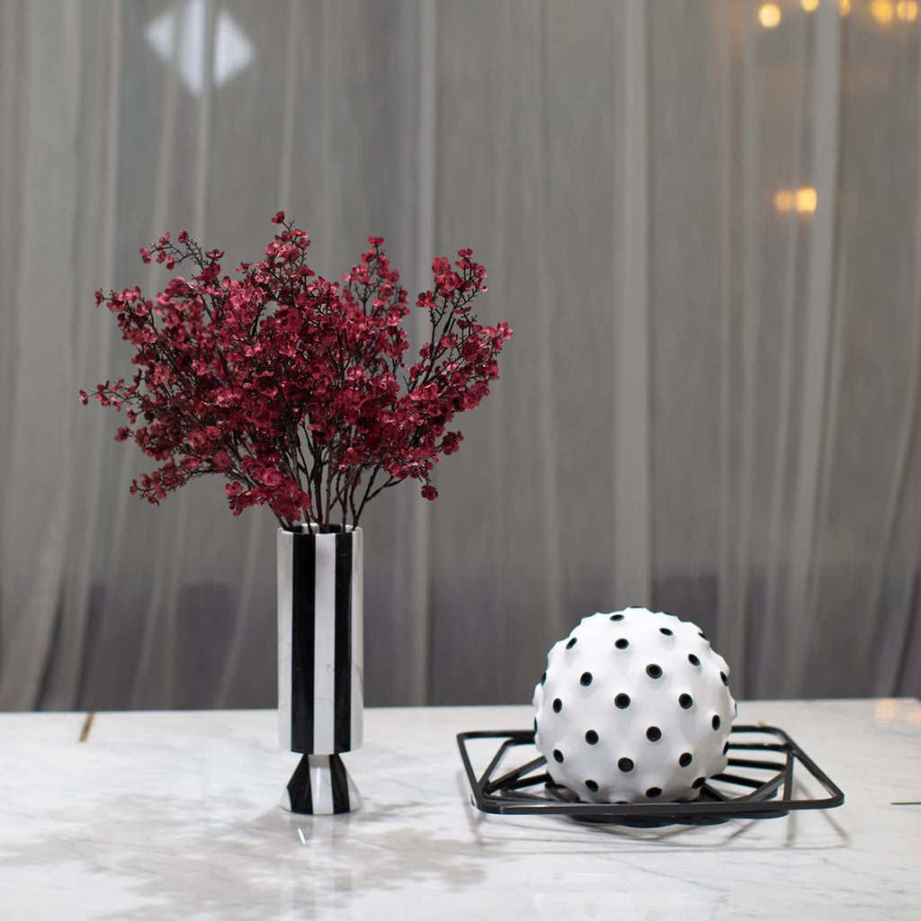 Burgundy artificial baby's breath flowers in a striped vase next to a decorative orb on a marble surface.