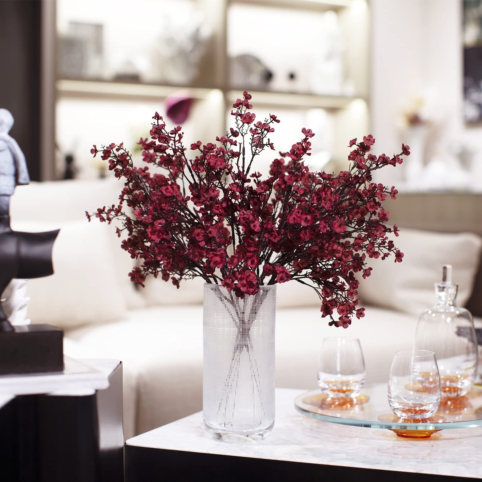 Burgundy artificial baby's breath flowers in a textured glass vase on a marble table with glasses and a decanter in the background.