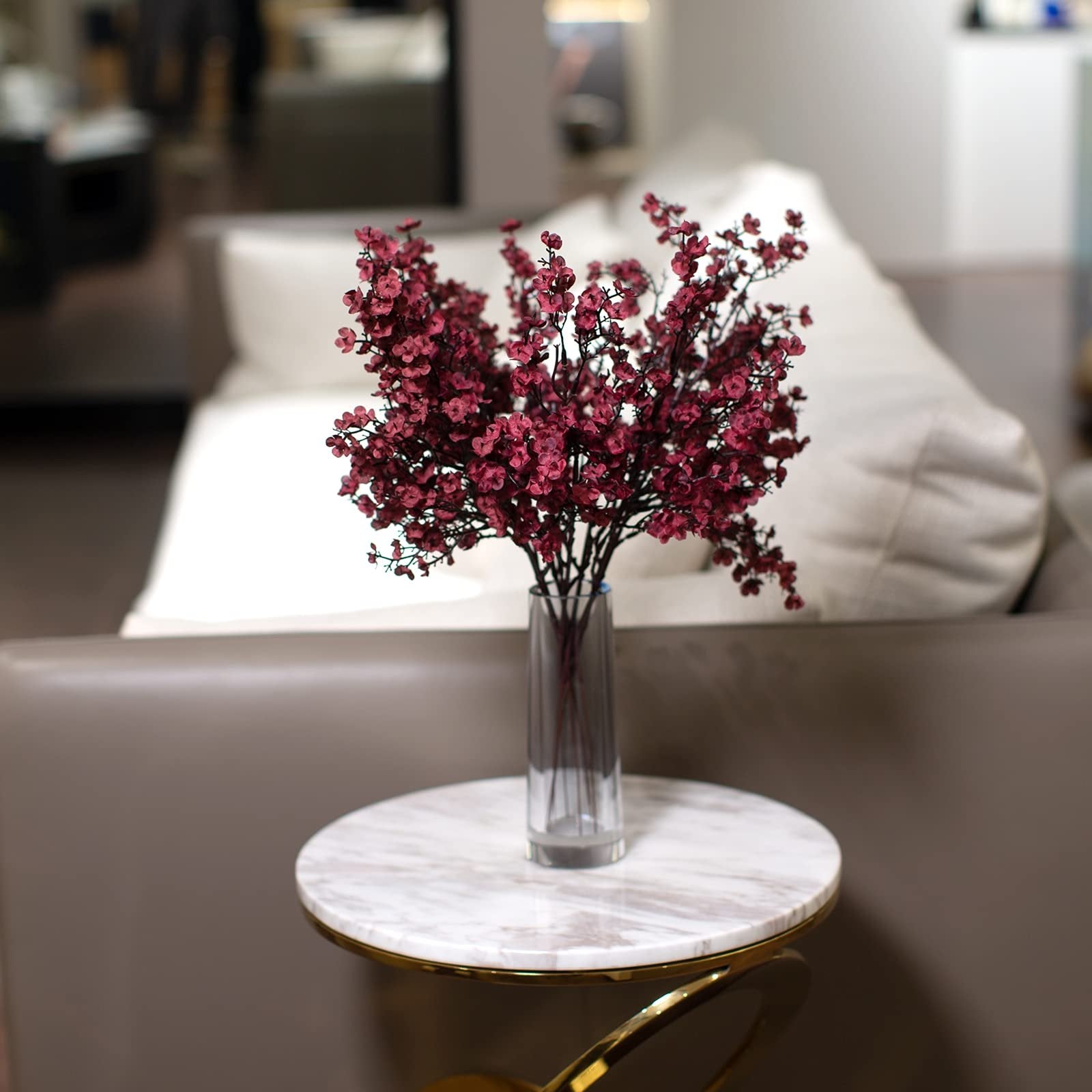 Burgundy artificial baby's breath flowers in a glass vase on a marble side table in a living room setting.