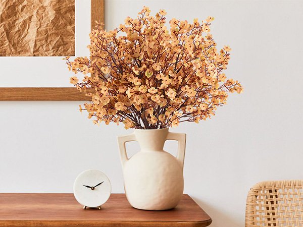 Artificial baby's breath flowers in a cream vase on a wooden desk with a small clock.
