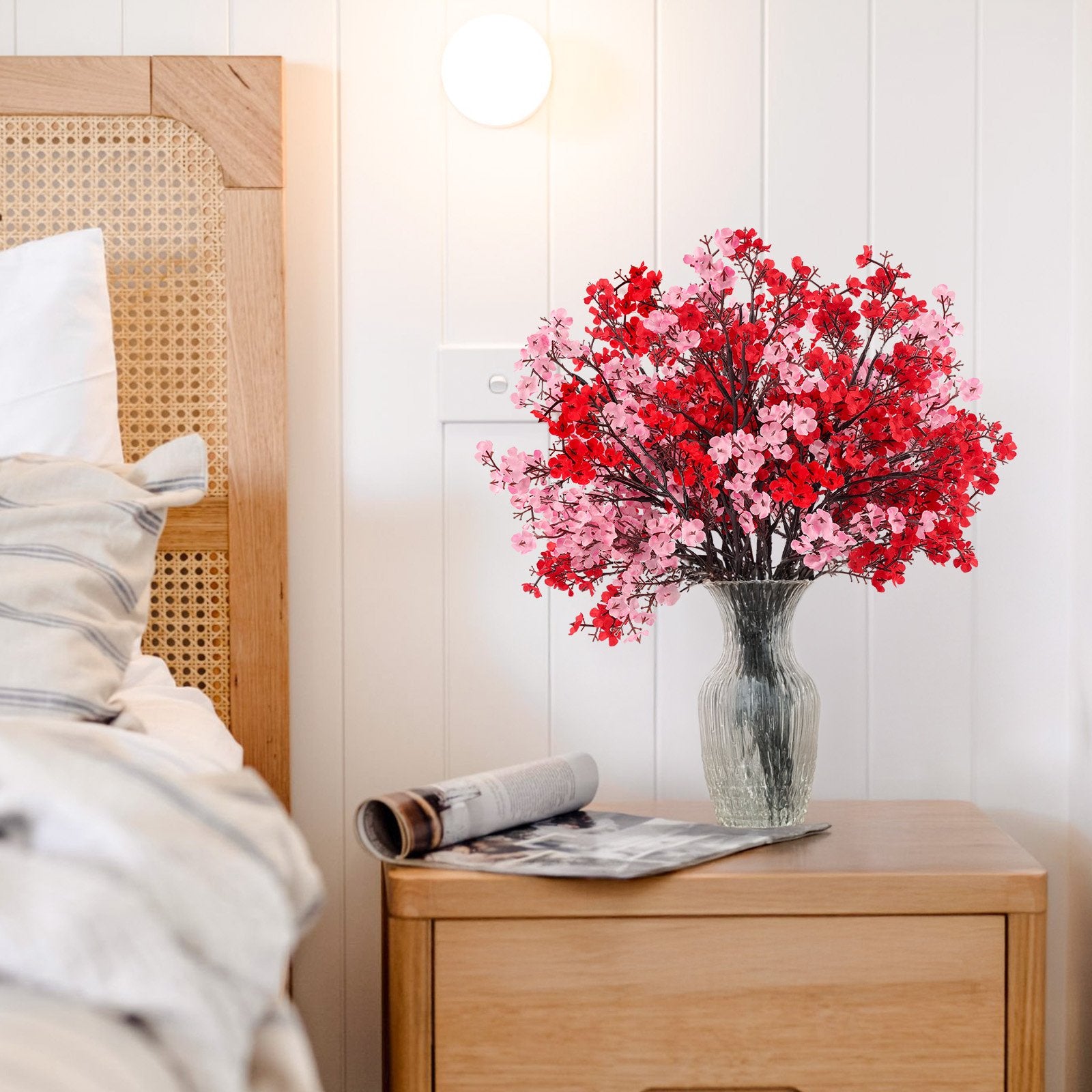 A vase of red and pink artificial baby's breath flowers on a wooden nightstand next to a bed.