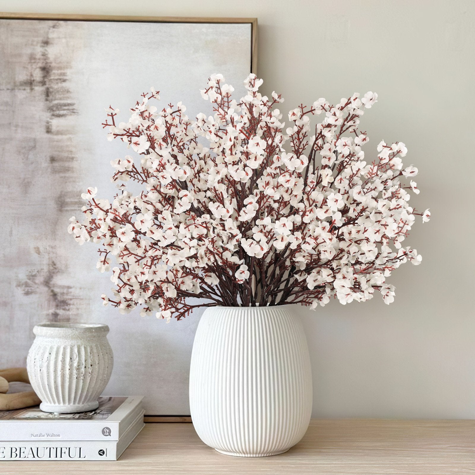 White baby's breath artificial flowers in a fluted vase on a wooden surface with books and a decorative pot.