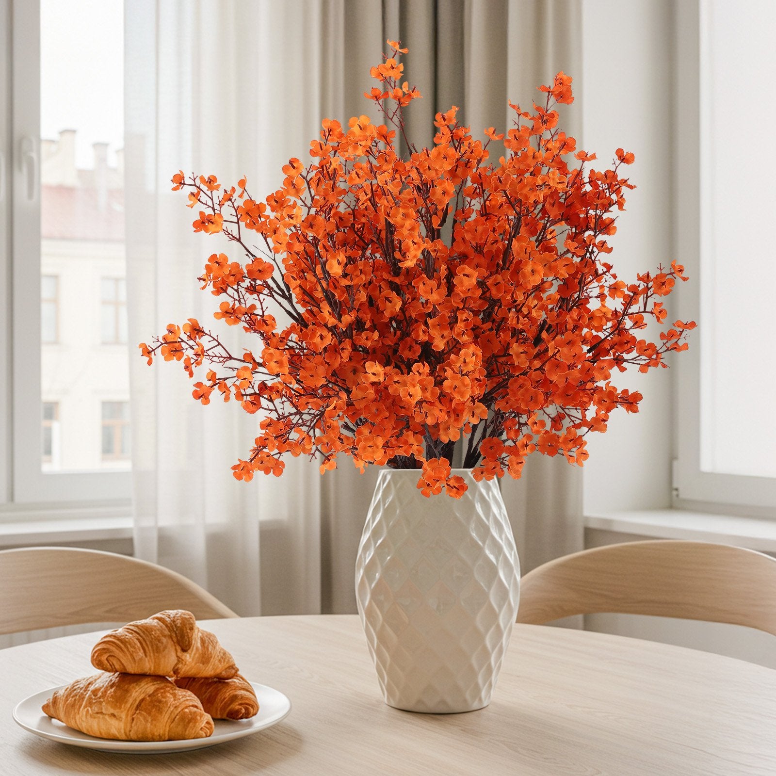Orange artificial baby's breath flowers in a white vase on a table with croissants