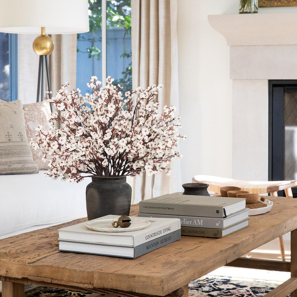 White baby's breath artificial flowers in a dark vase on a rustic wooden coffee table with books and decor.