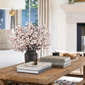 White baby's breath artificial flowers in a dark vase on a rustic wooden coffee table with books and decor.