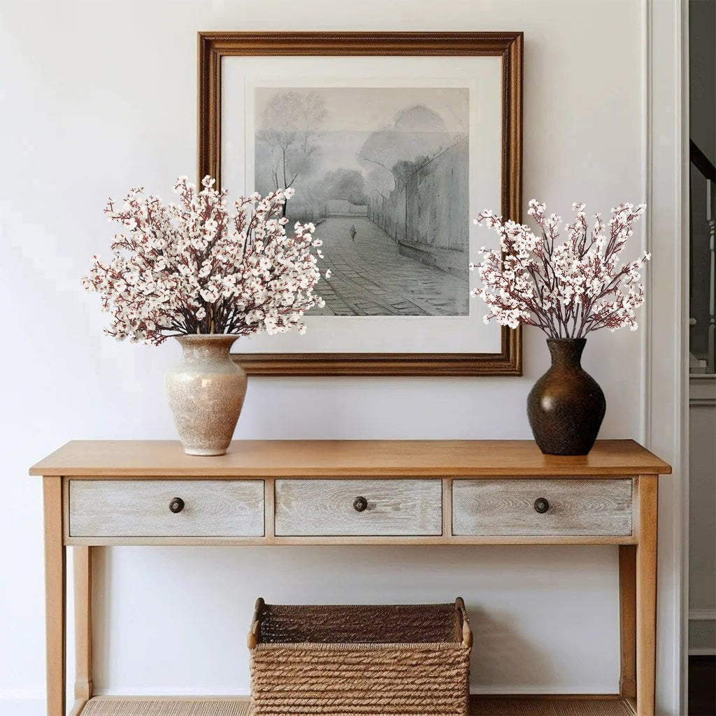 Two vases of white baby's breath artificial flowers on a wooden console table in front of a framed picture.