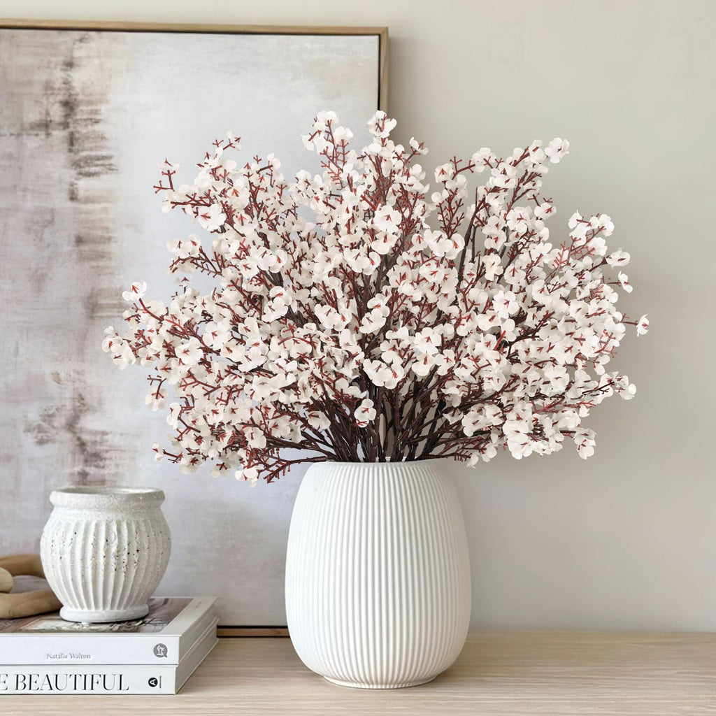 White baby's breath artificial flowers in a fluted white vase on a wooden surface with books and a textured vase.