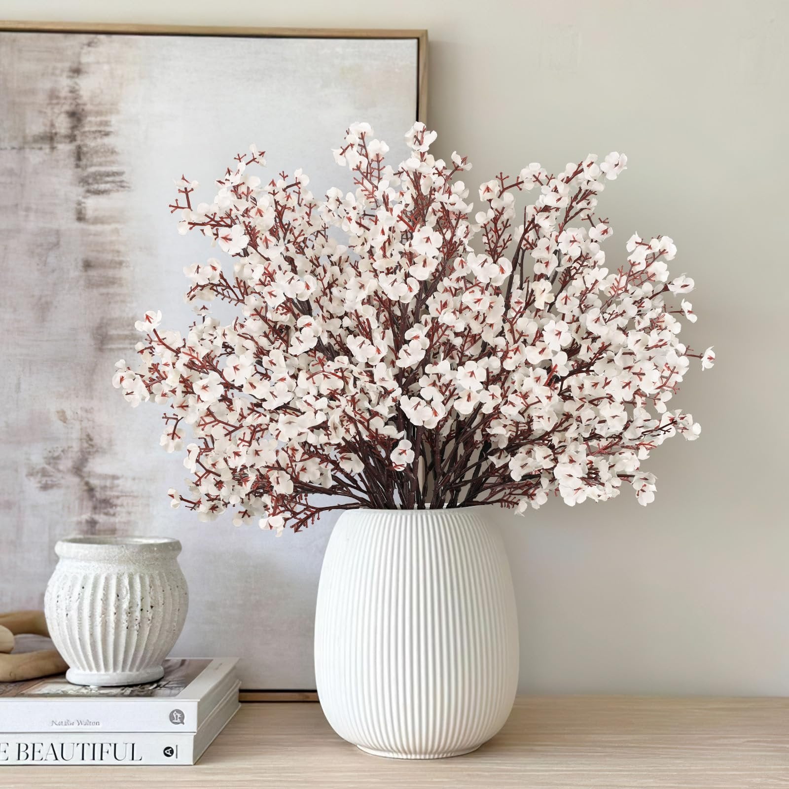 White baby's breath artificial flowers in a fluted white vase on a wooden surface with books and a textured vase.