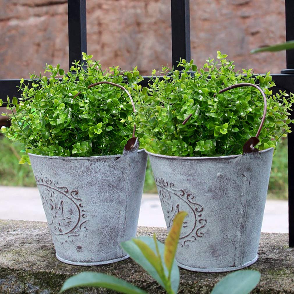 Two distressed metal buckets filled with artificial boxwood greenery, set on a stone ledge with a black metal fence behind.