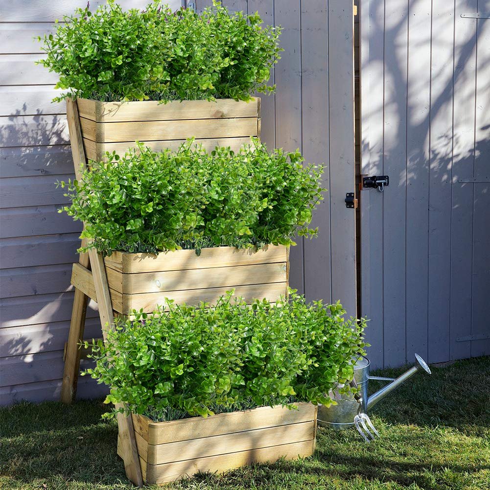 Tiered wooden planter filled with artificial boxwood stems on a grassy lawn next to a wooden gate and watering can.