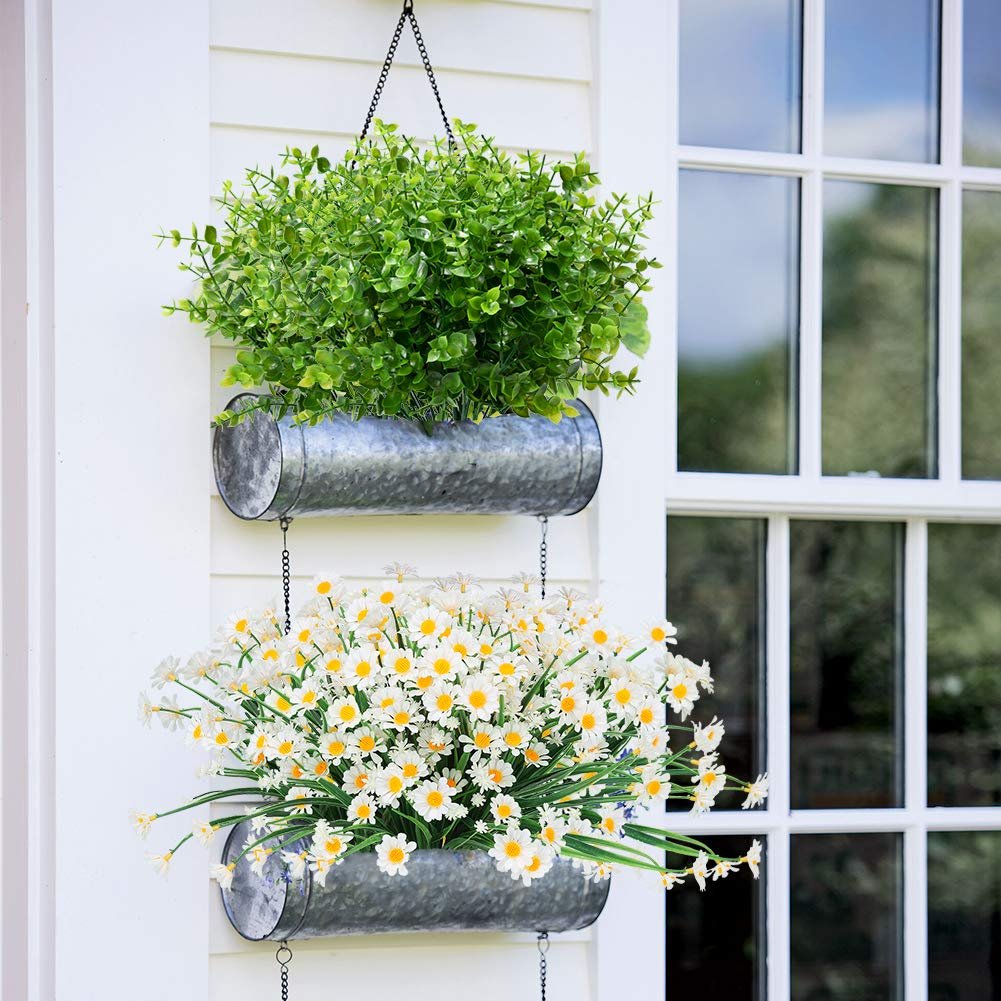 Two galvanized planters hanging on a white wall next to a window, one with green boxwood stems and the other with white daisies.