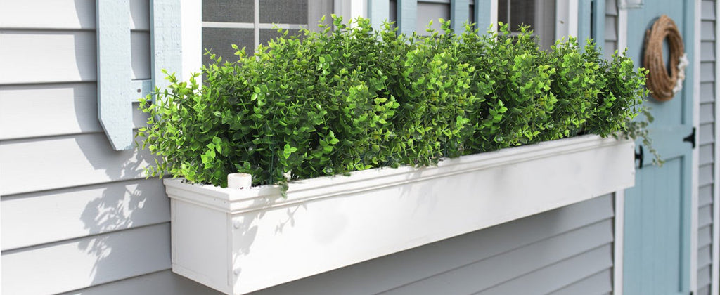 Artificial eucalyptus greenery in a white window box planter mounted on a house exterior with blue shutters and a wreath.