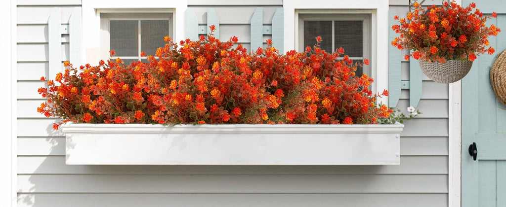 Artificial fall flowers with orange and red blooms in a white window box and hanging basket on a house exterior.