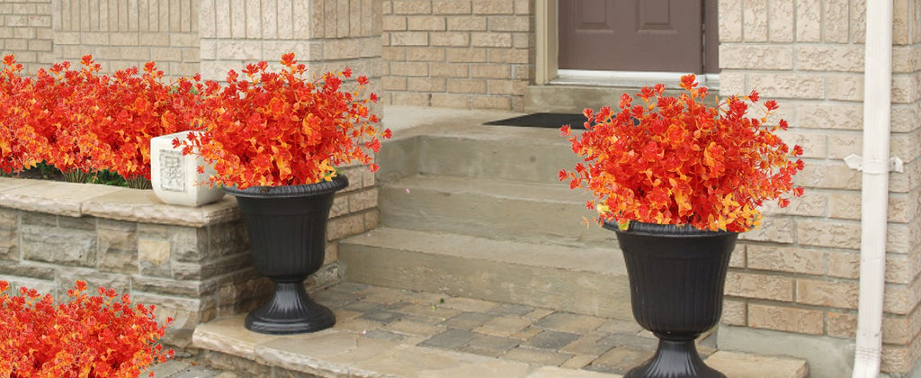 Two black urn planters filled with vibrant orange and red artificial fall foliage plants on a stone porch with brick walls.