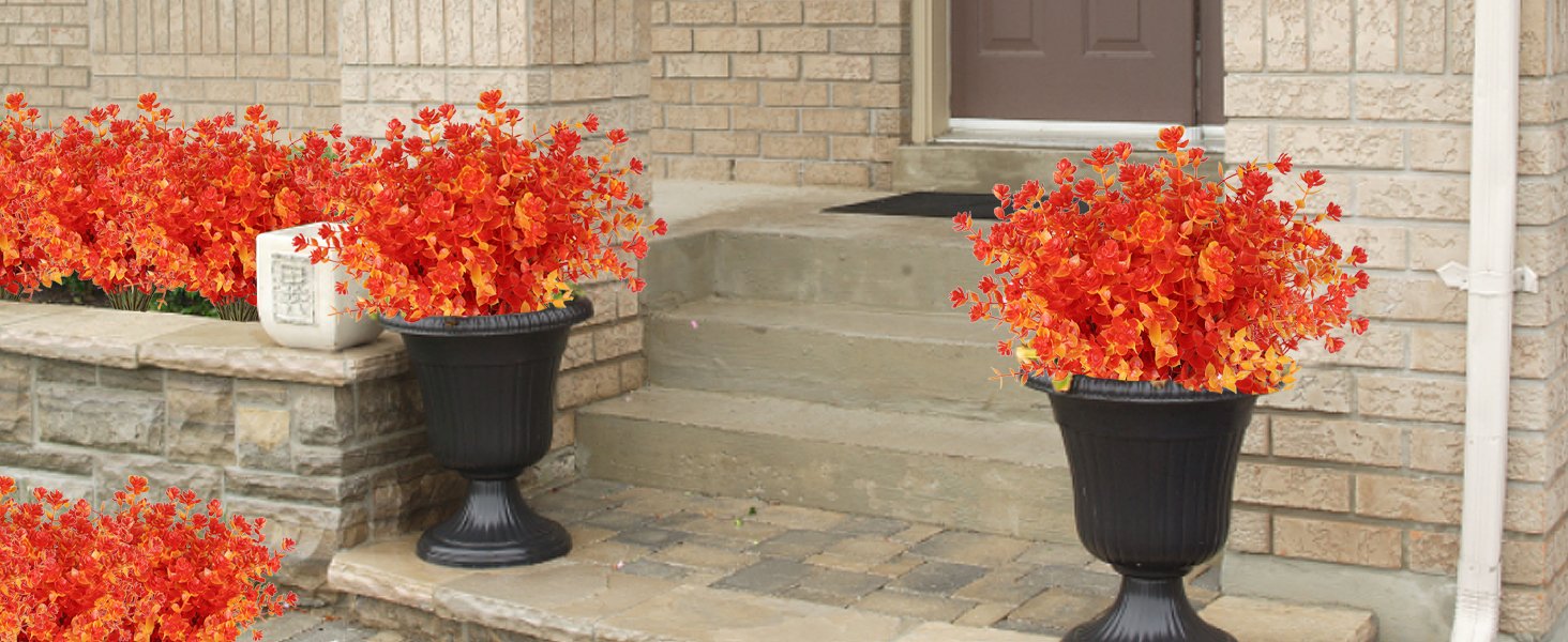 Two black urn planters filled with vibrant orange and red artificial fall foliage plants on a stone porch with brick walls.
