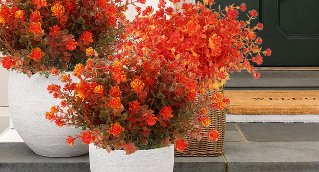 Artificial fall flowers in orange and red hues with green leaves in white planters and a wicker basket near a doorway.