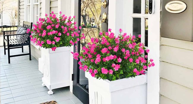 Two white planters filled with vibrant pink artificial flowers and green foliage on a tiled porch next to a doorway.