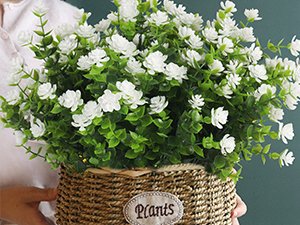White artificial flowers with green eucalyptus leaves in a woven basket, held by hands.