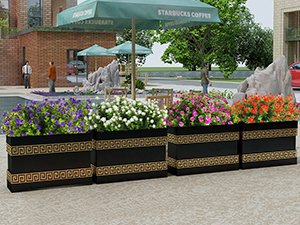 Four black planters with gold Greek key trim filled with colorful artificial flowers, set outdoors near a building with umbrellas.