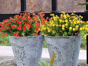 Two decorative metal buckets filled with artificial red and yellow flowers, placed outdoors near a fence.