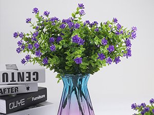Artificial purple flowers with green leaves in a blue and pink vase, with books in the background.