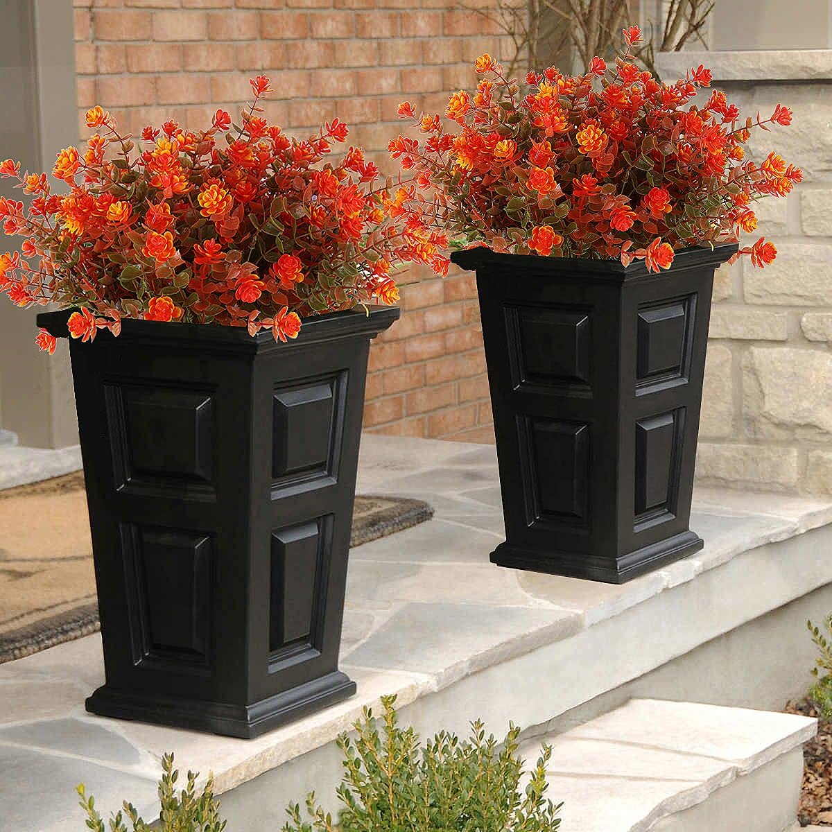 Two black planters filled with artificial orange and red fall flowers on a stone porch.