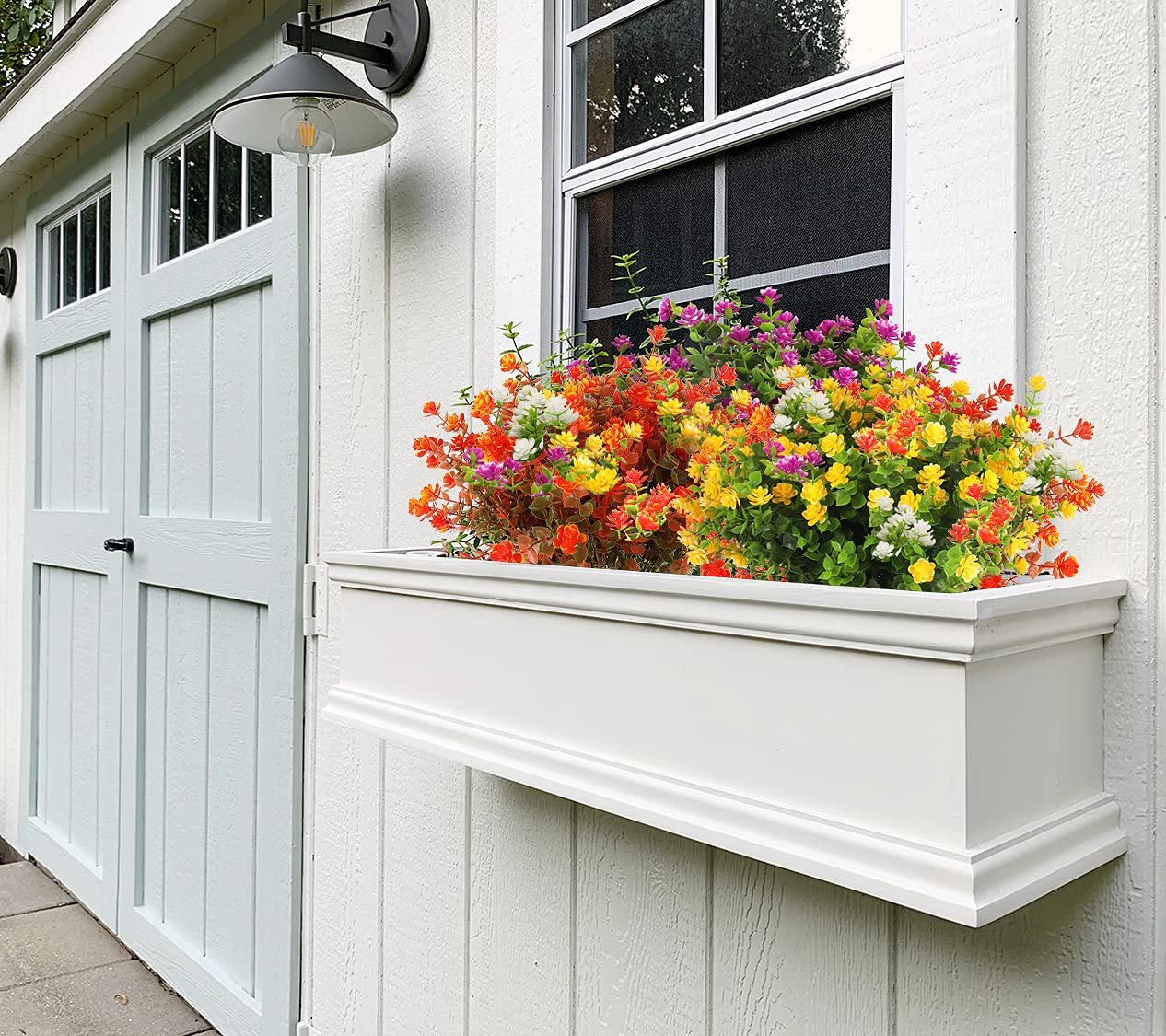 Window box filled with colorful artificial fall flowers and eucalyptus leaves mounted on a white shed wall.