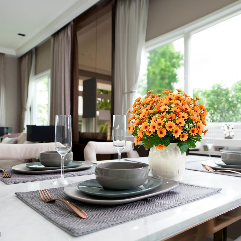 Arrangement of orange artificial sunflowers in a white vase on a dining table setting.