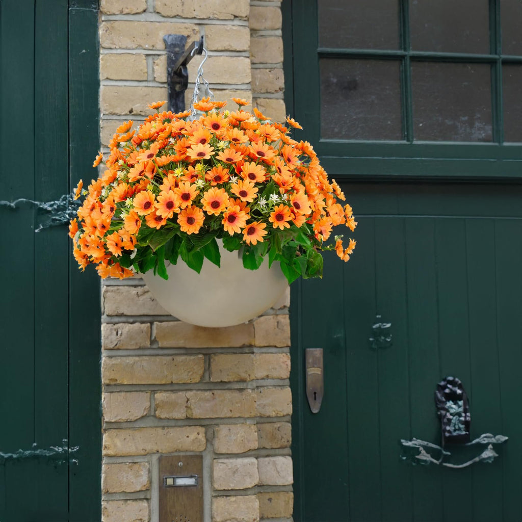 Hanging basket filled with vibrant orange artificial sunflowers and green leaves, suspended from a hook on a brick wall next to a dark green door.