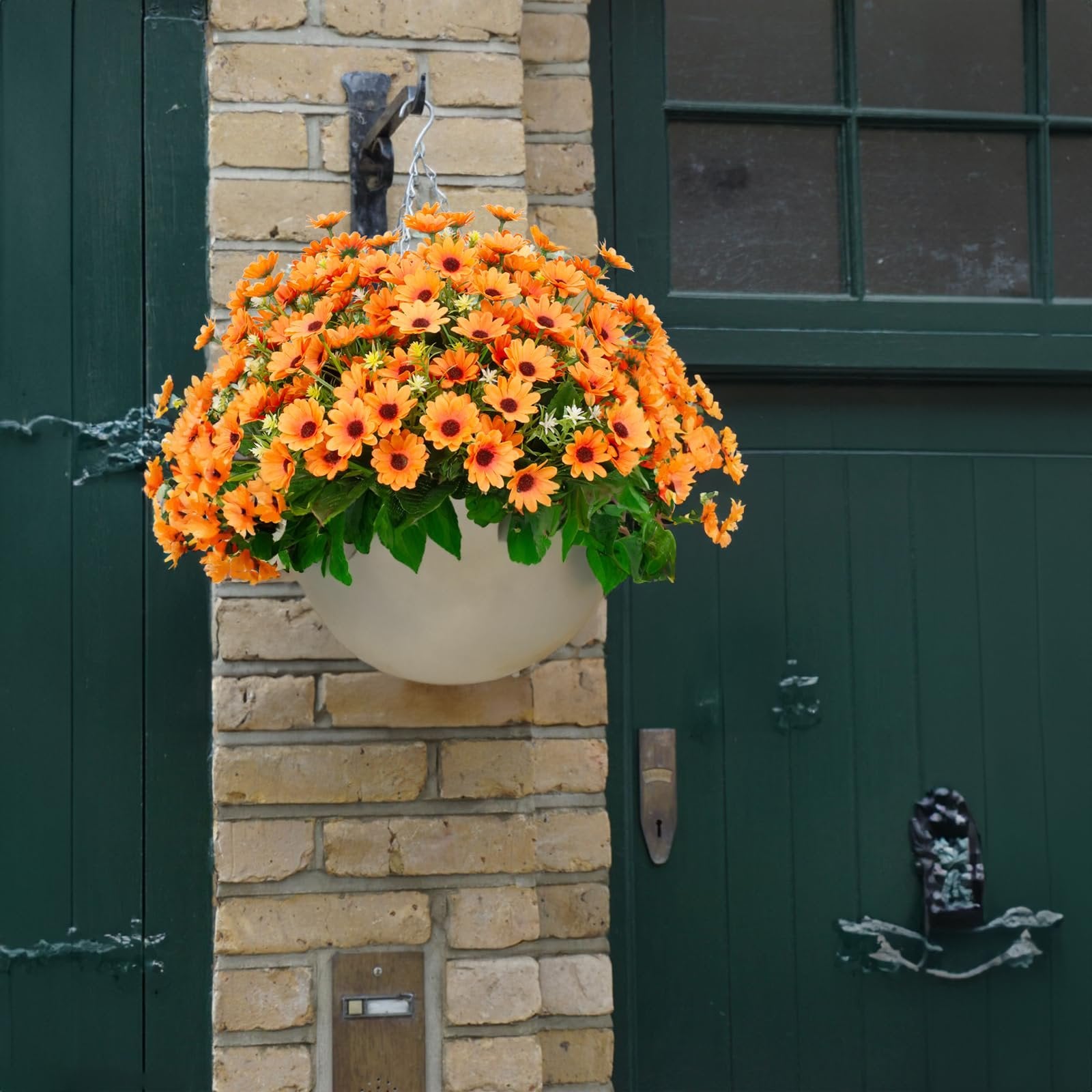 Hanging basket filled with vibrant orange artificial sunflowers and green leaves, suspended from a hook on a brick wall next to a dark green door.