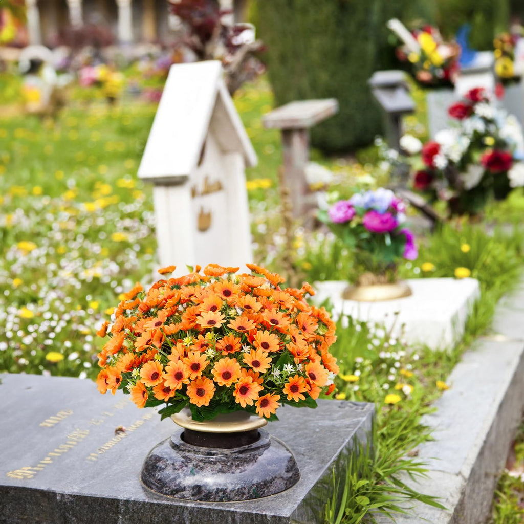 Arrangement of orange artificial daisies in a vase on a tombstone in a cemetery with other flowers and grave markers in the background.