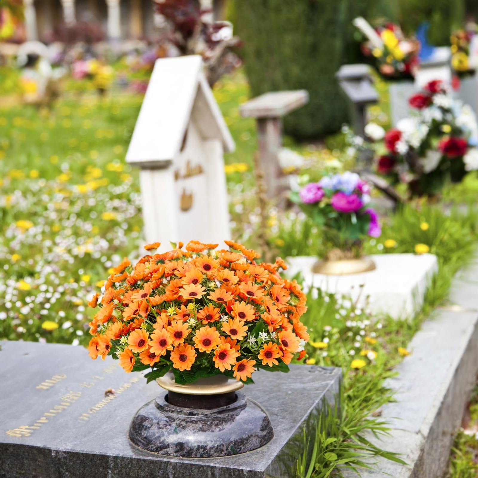 Arrangement of orange artificial daisies in a vase on a tombstone in a cemetery with other flowers and grave markers in the background.