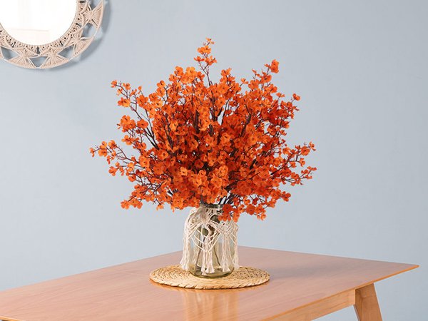 Eight bundles of orange artificial gypsophila flowers in a glass vase on a wooden table, with a macrame detail on the vase and a decorative mirror in the background.