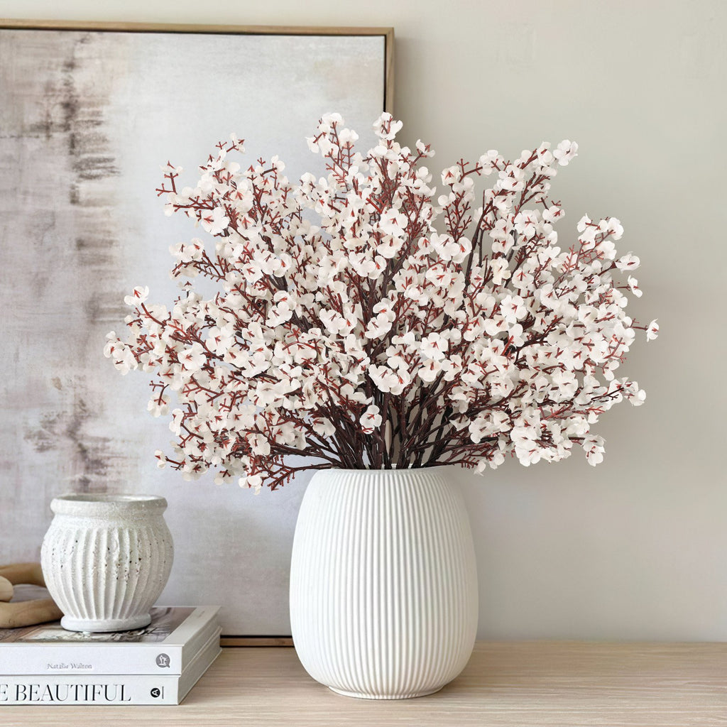 Eight bundles of light brown artificial gypsophila flowers in a white ribbed vase on a wooden surface, with books and a decorative pot nearby.