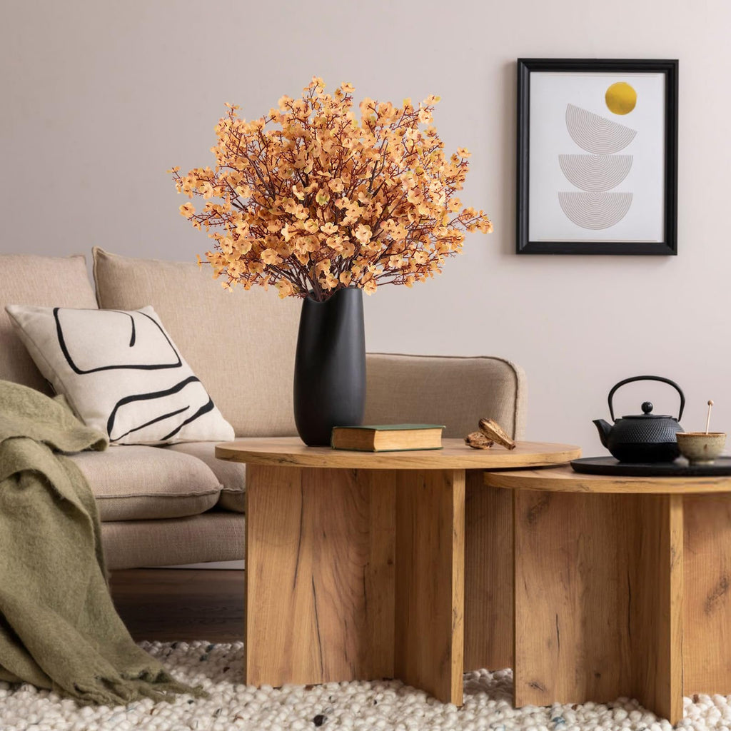 Light brown artificial gypsophila flowers in a black vase on a wooden coffee table, next to a sofa and framed art.