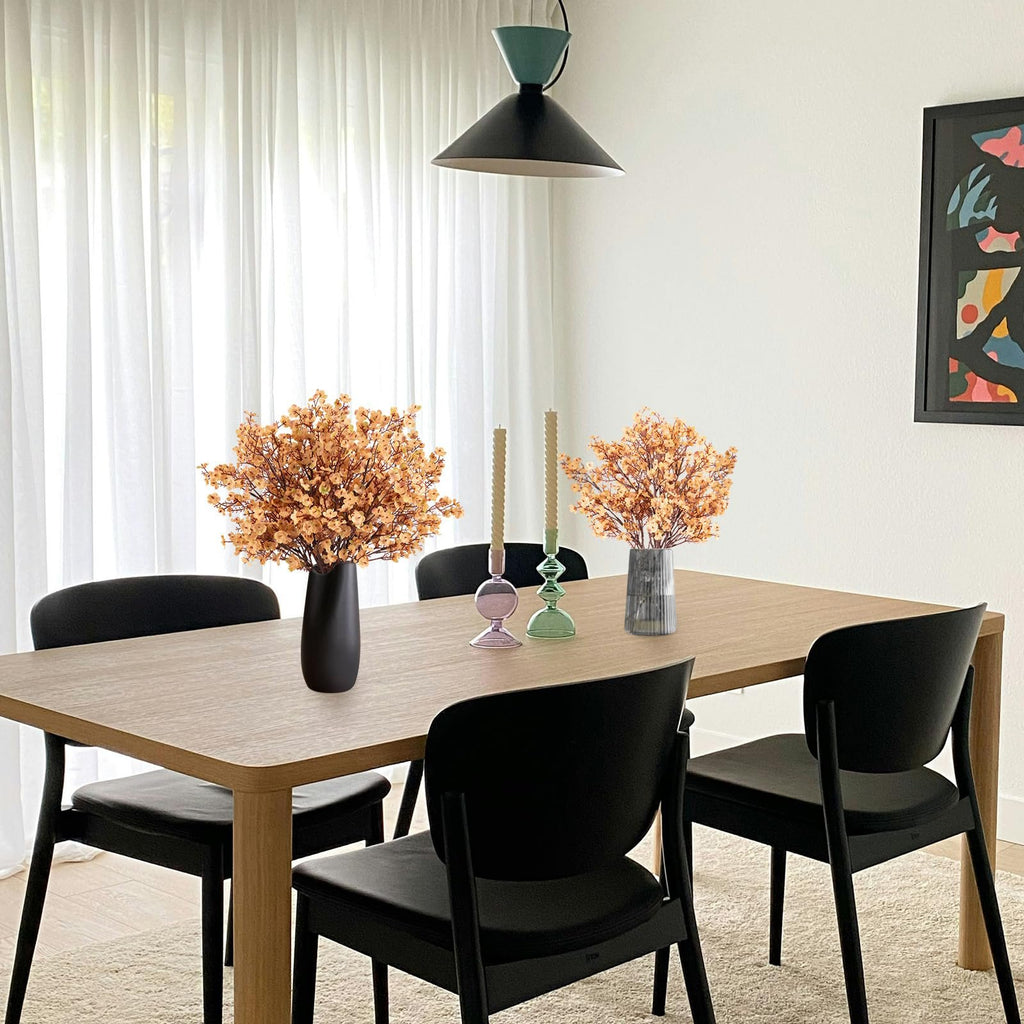 Dining table set with two vases of light brown artificial flowers, two candles, and black chairs.