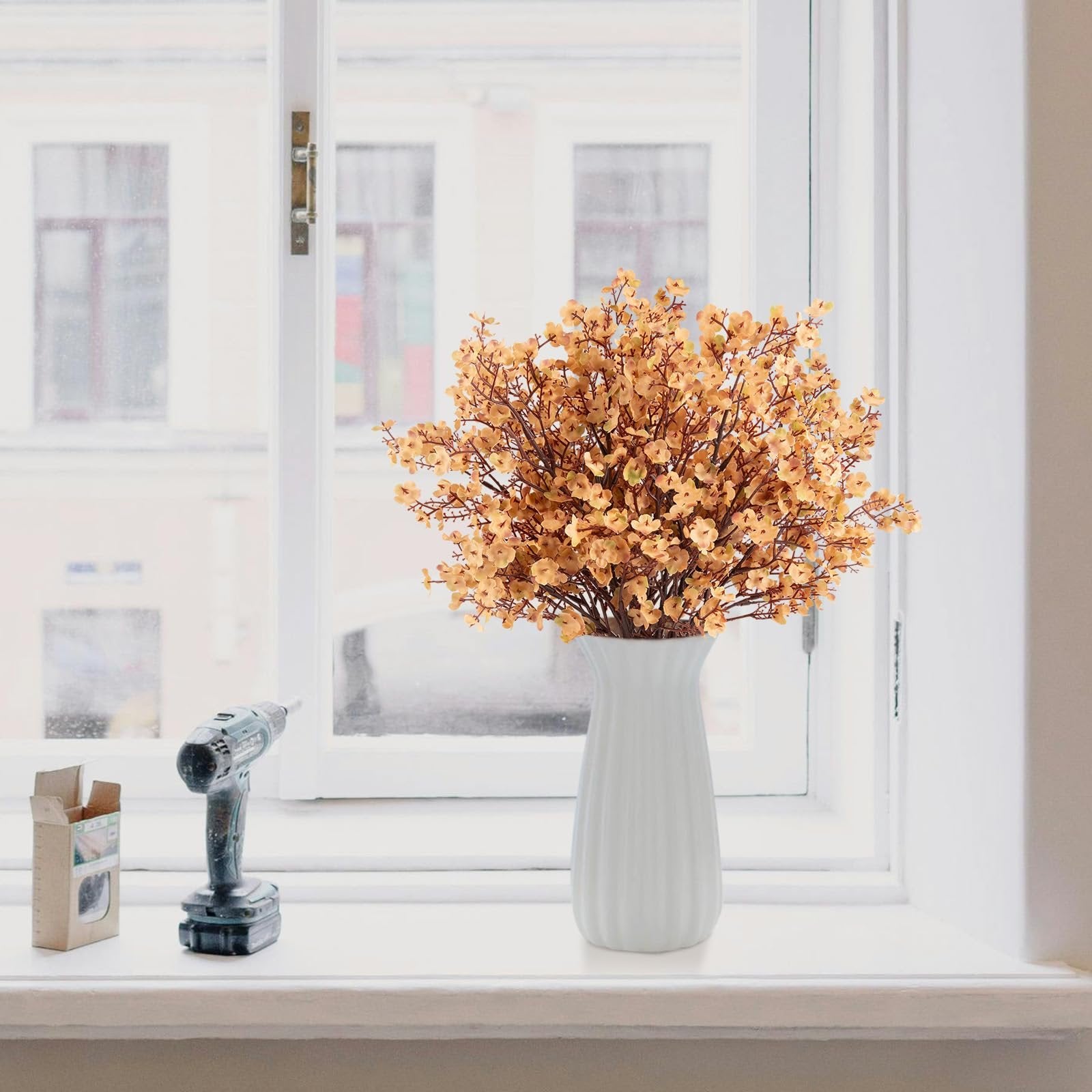 Light brown artificial gypsophila flowers in a white vase on a windowsill next to a drill and box.