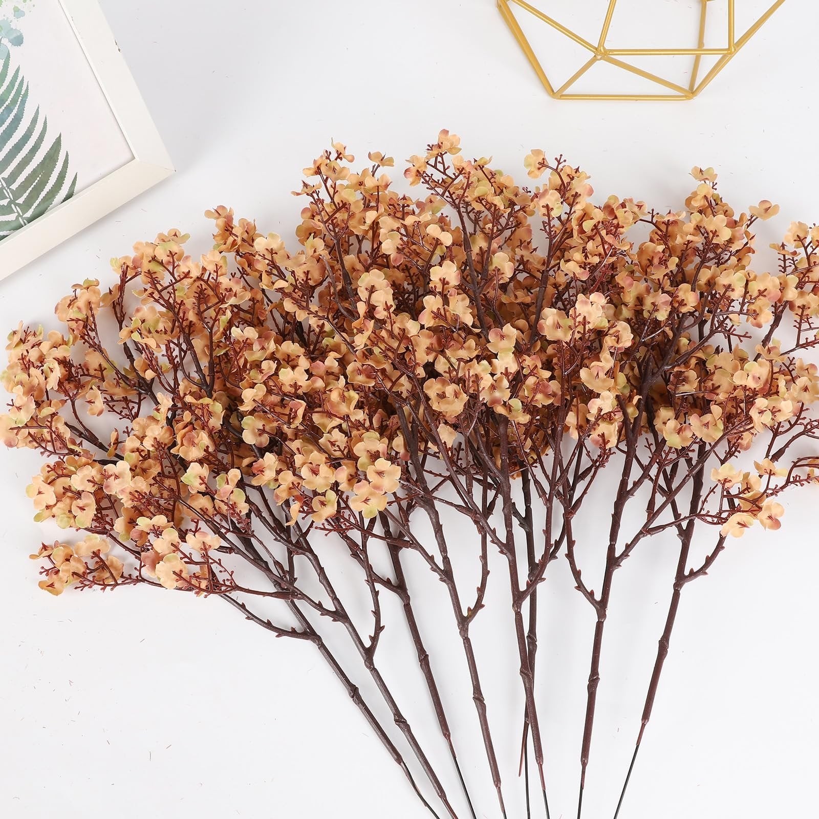 Eight bundles of light brown artificial gypsophila flowers with delicate petals and brown stems, arranged on a white background with a framed picture and gold geometric decor.
