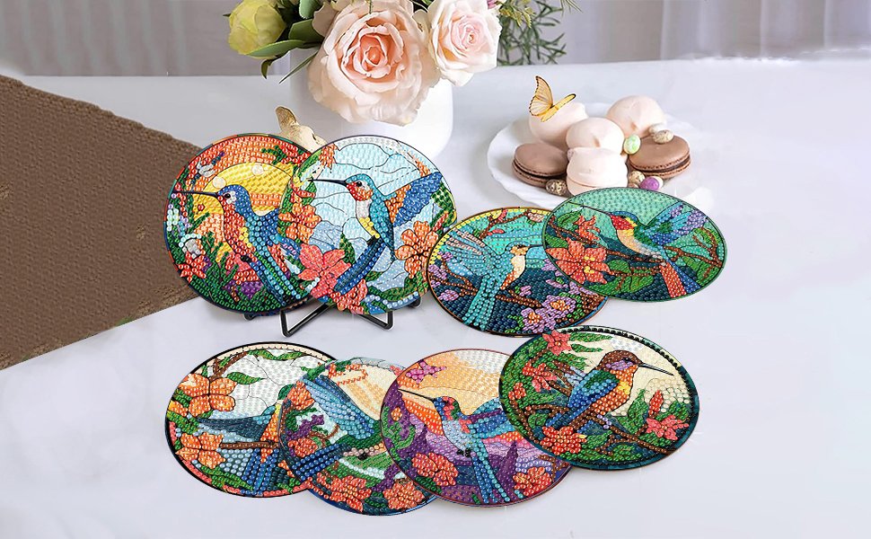 Eight round diamond painting hummingbird coasters with holder, displayed on a table with flowers and macarons.