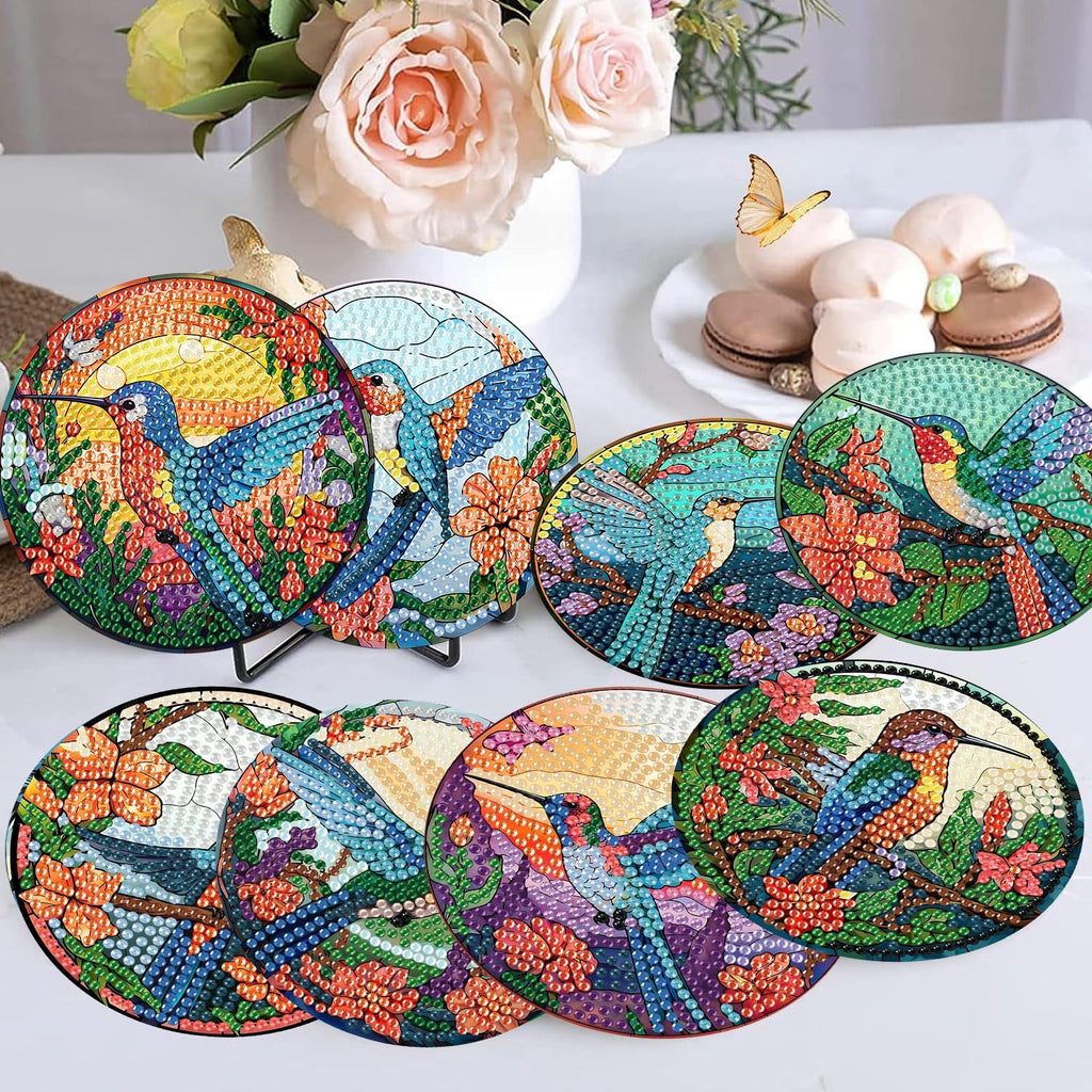 Eight diamond painting hummingbird coasters with holder, displayed on a white surface with flowers and macarons in the background.