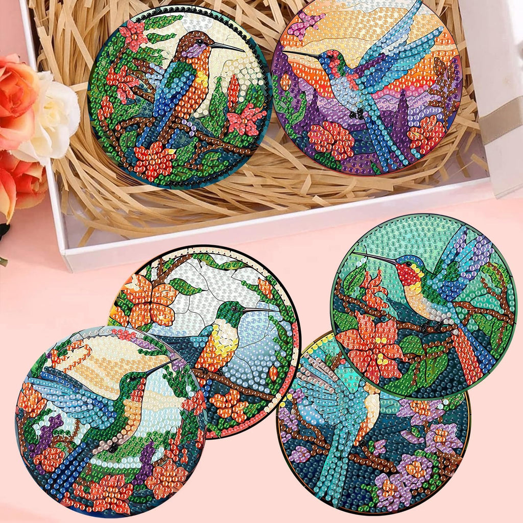 Diamond painting coasters with hummingbird designs and holder, arranged in a white box with straw filler and decorative flowers.