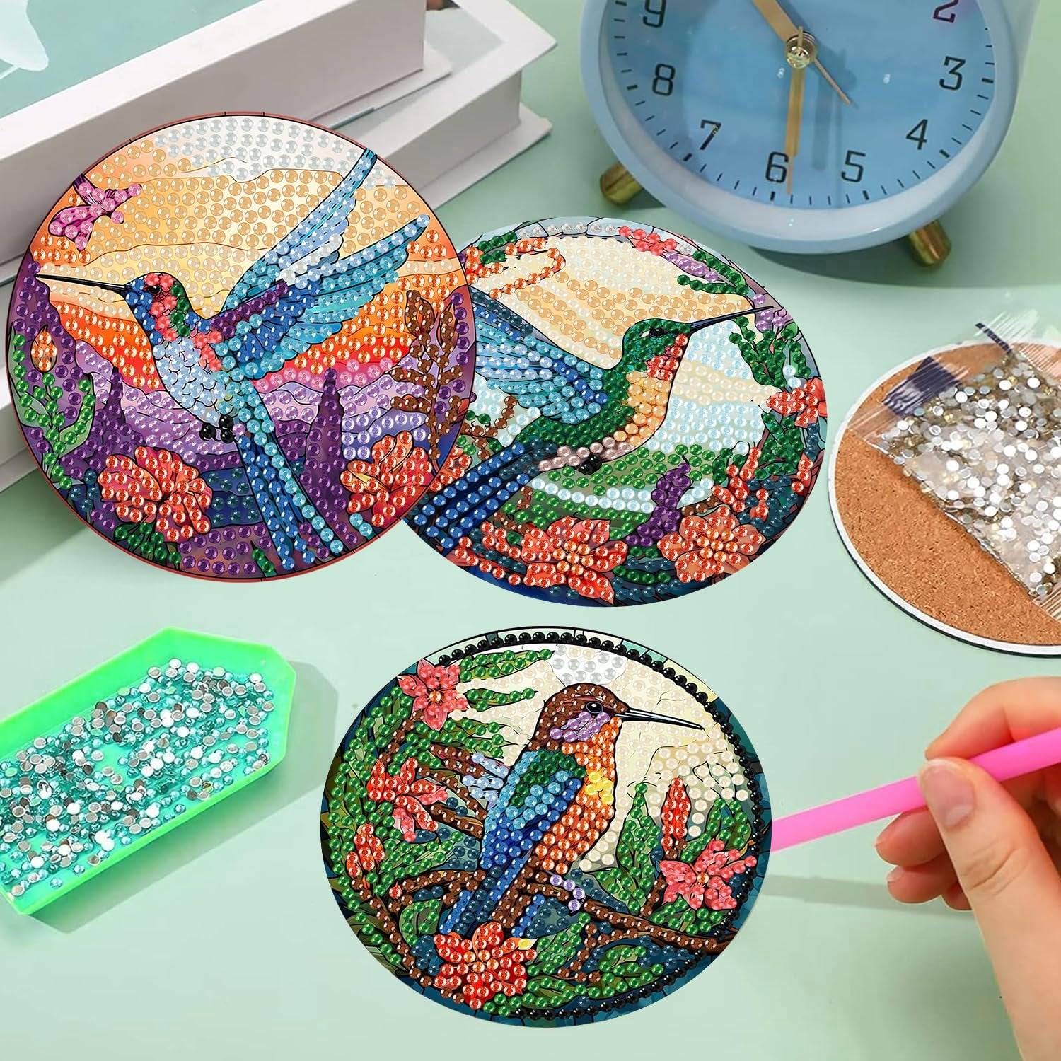Diamond painting coasters with hummingbird designs, tray of gems, and a pink tool