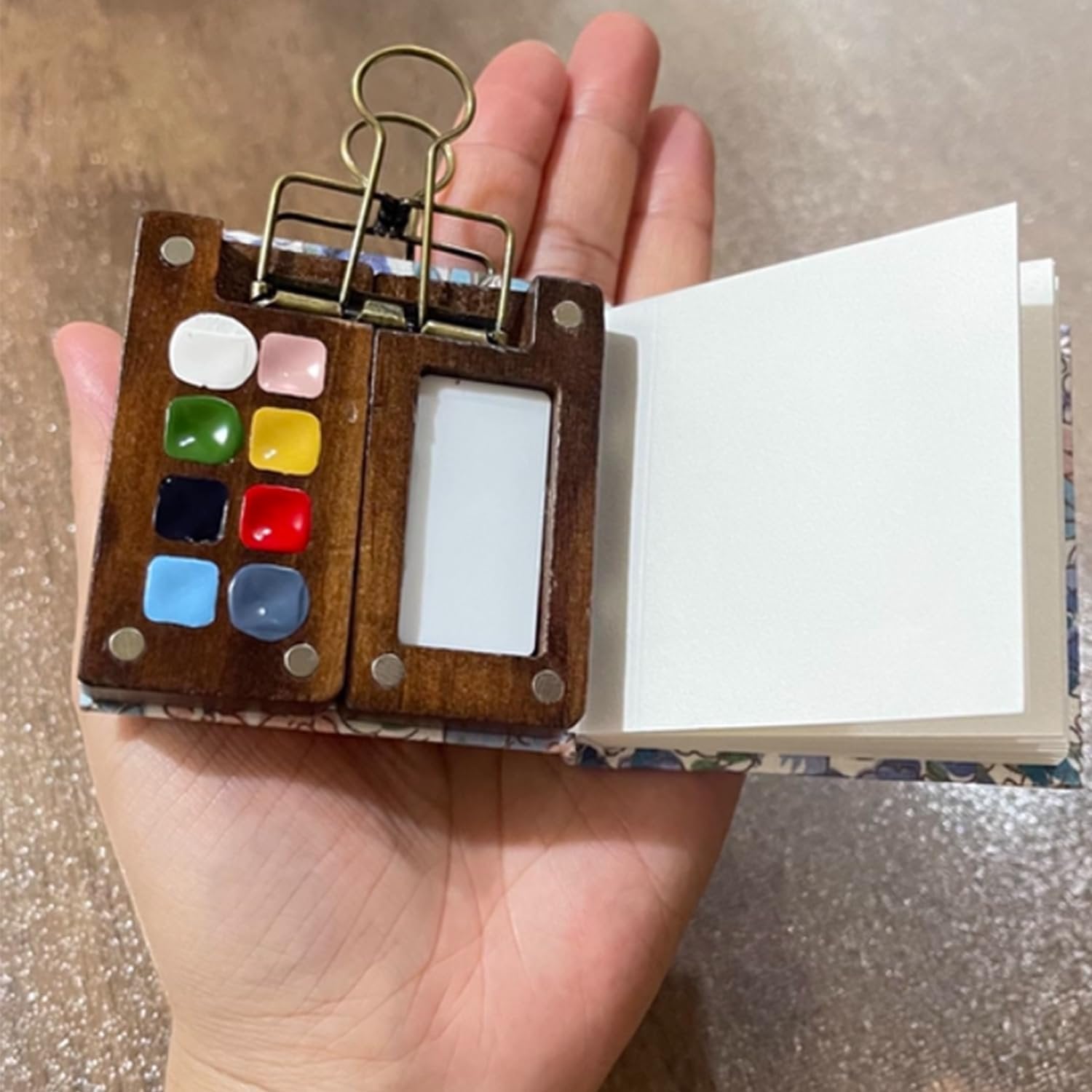 Hand holding a mini watercolor paint set with 8 colors and a small sketchbook, open to blank pages.