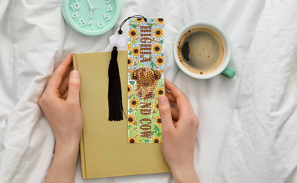 Diamond art bookmark with sunflowers and a highland cow design, held by hands next to a book and coffee cup.