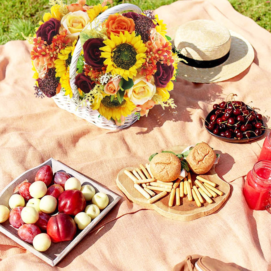 Artificial sunflower and rose bouquet in a white basket on a picnic blanket with fruit and snacks