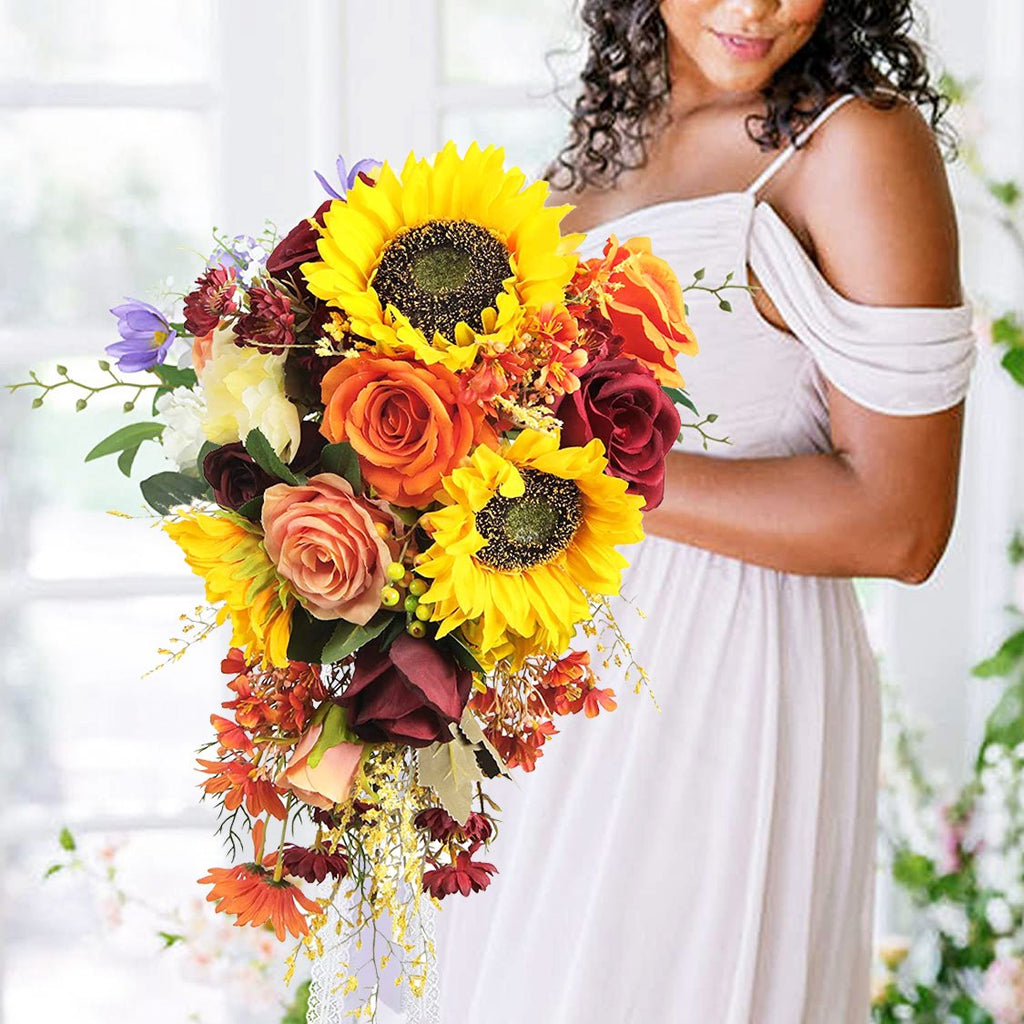 Woman in white dress holding a cascading bouquet of artificial sunflowers, roses, and other fall-colored flowers.